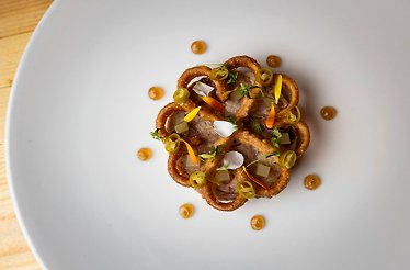 Overhead view of a fine dining dish with crispy fried onion rings, sliced jalapeños, edible flowers, herbs, and golden sauce dots.