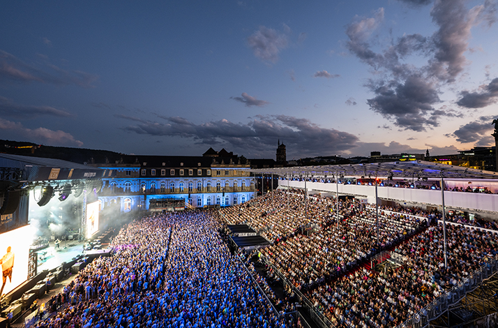 Aerial view of Jazz Open Stuttgart at twilight, thousands of attendees filling the courtyard of a historic illuminated palace.
