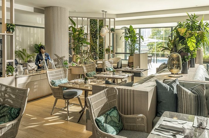 Sunlit Four Seasons restaurant with wicker chairs, lush plants, and tables set beside large windows.