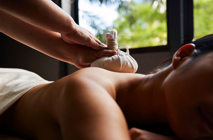 Therapist applying a herbal compress massage to a guest’s back near a window at the spa.