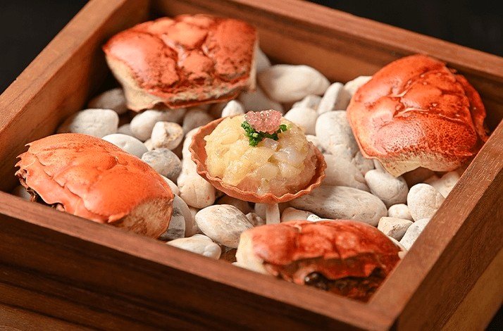 A dish of crab meat in a shell is presented in a wooden box on a bed of white stones.