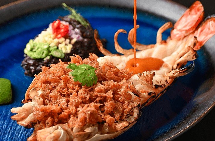 A dish of a large, cooked crustacean with black rice and topped with a sauce.