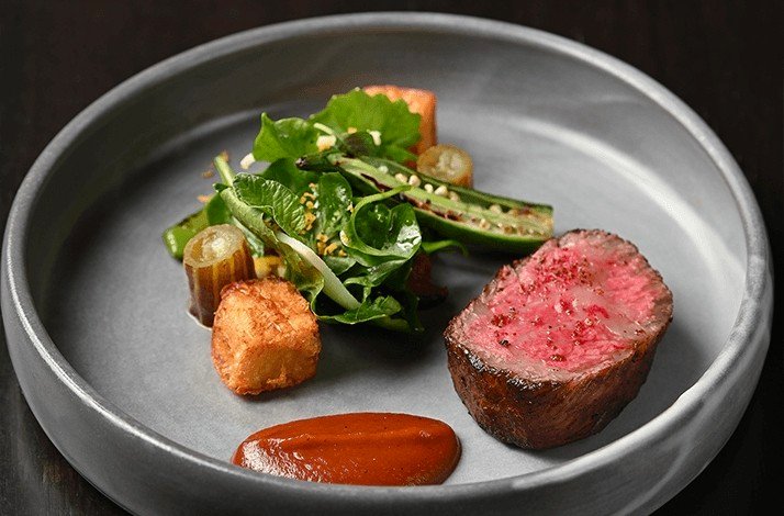 A dish of medium-rare steak is served with a side of mixed greens and a red sauce.