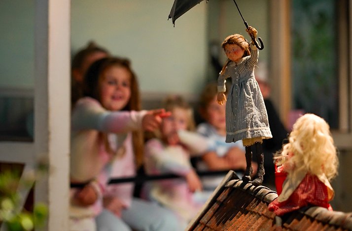 A small doll holding an umbrella stands on a rooftop in front of seated figures.