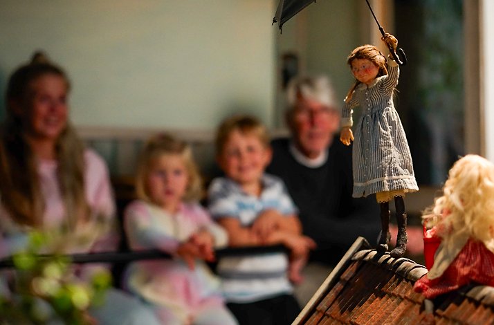 A small doll holding an umbrella stands on a rooftop in front of seated figures.