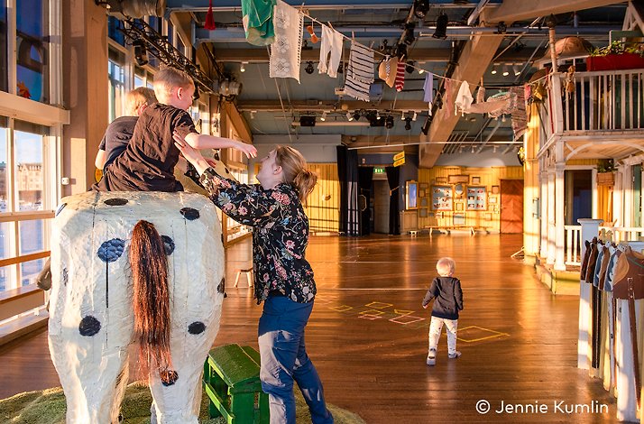 Children playing indoors; one climbs a large spotted cow sculpture. © Jennie Kumlin