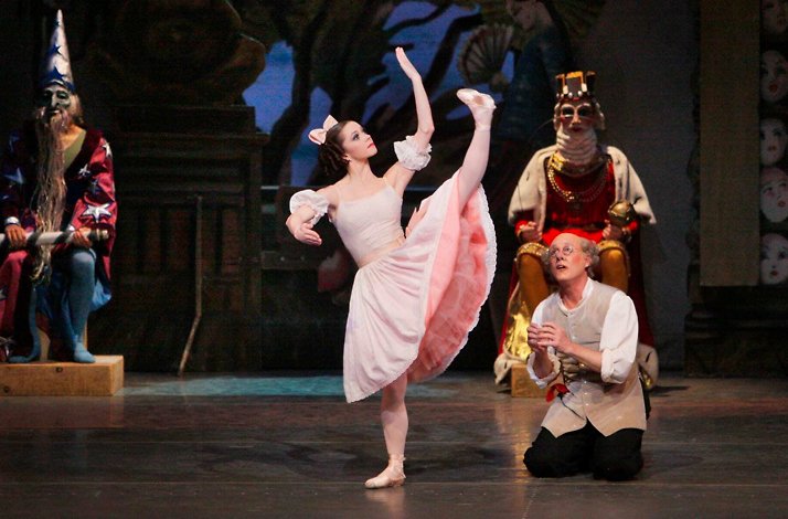 Ballet dancer in pink costume performs on stage with theatrical set and seated characters.