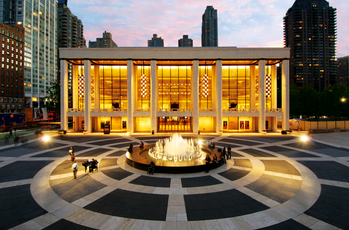 Exterior of Lincoln Center at dusk, glowing lights and fountain in front, New York City skyline behind.