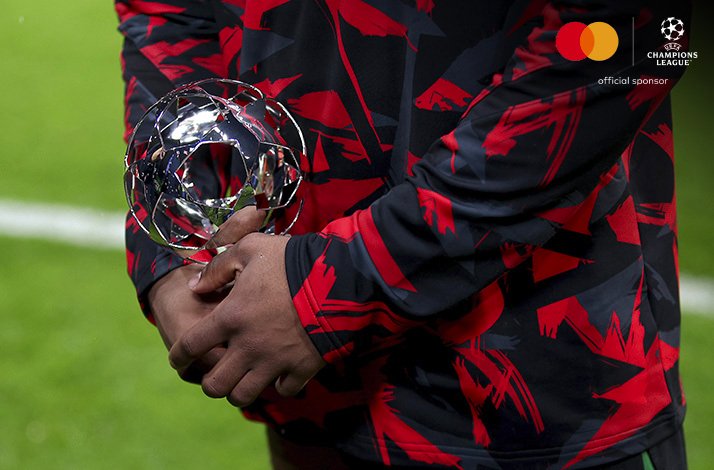 Player holding a UEFA Champions League star-ball award trophy, wearing a red and black warm-up jacket.