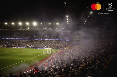 Packed stadium during a UEFA Champions League match, with fans and smoke filling the stands under bright lights.