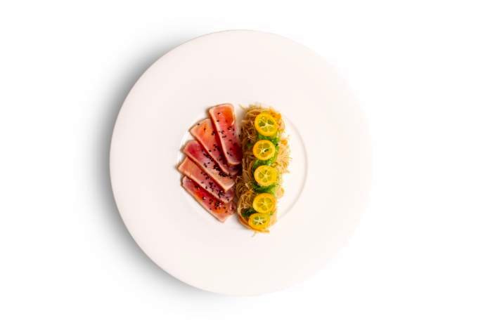 Seared sesame-crusted tuna slices paired with thin noodles and bright sliced kumquats on a white plate.