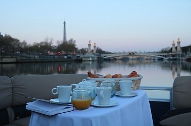 View from aboard cruise boat overlooking Seine and Eiffel Tower. On boat there is table with teapot, tea cups and croissants prepared for breakfast. 