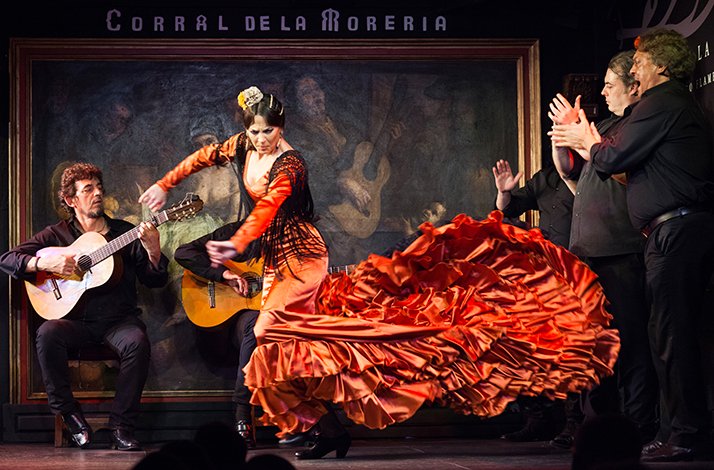 The performance of flamenco dance on stage of Corral de la Moreria. Woman is dancing in traditional red dress, and 2 men are playing guitars.