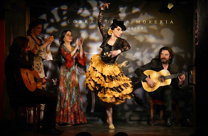 The performance of flamenco dance on stage of Corral de la Moreria. Woman is dancing in traditional yellow skirt among 2 women, and 2 men are playing guitars.