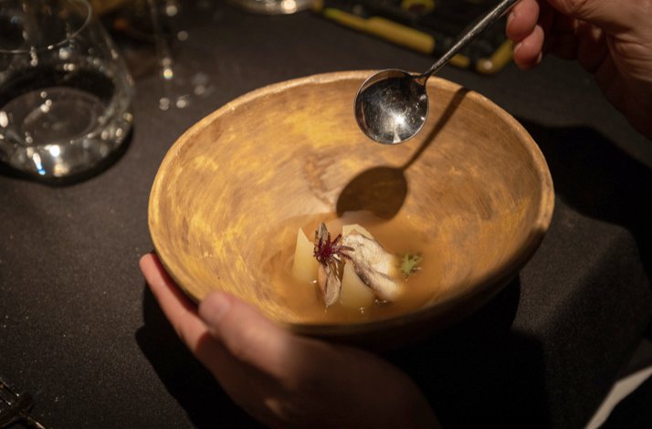 A person holds a rustic wooden bowl with a gourmet dish, including fish, herbs, and broth. 
