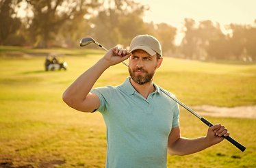 A man is standing on a sunny golf field, holding his hand by the cap visor.