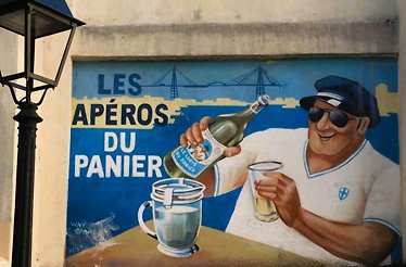 Street art in Le Panier, Marseille's oldest district - an image on the building wall displaying a man enjoying a local drink with the words reading “LES APEROS DU PANIER”.