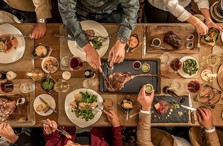 Overhead view of diners sharing a tomahawk steak, salmon, and sides at Little London Bar and Grille, with wine glasses on a wooden table.