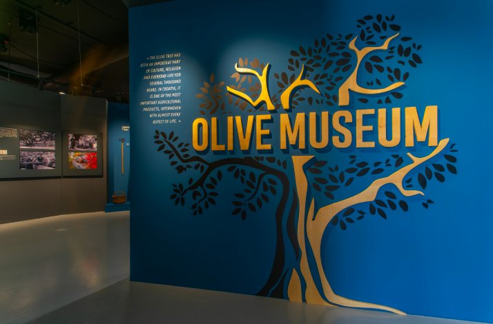 Golden and black picture of a tree with ‘OLIVE MUSEUM’ sign on a blue wall in Olive Museum.