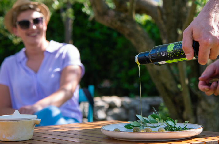 Person is pouring fresh olive oil onto a plate of olives and herbs during an outdoor tasting in a sunlit garden.