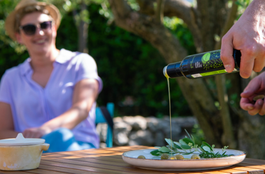 Person is pouring fresh olive oil onto a plate of olives and herbs during an outdoor tasting in a sunlit garden.