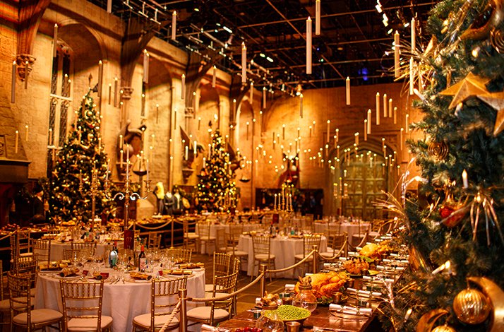 Candlelit banquet tables in a grand hall, with decorated Christmas trees, hanging candles, and festive place settings. 