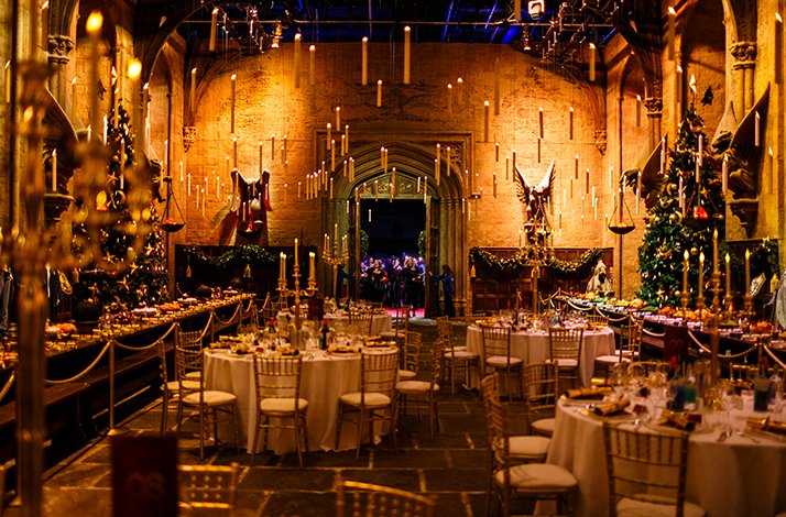 Festive dining hall with round tables, gold chairs, Christmas trees, and floating candles leading to an arched doorway.