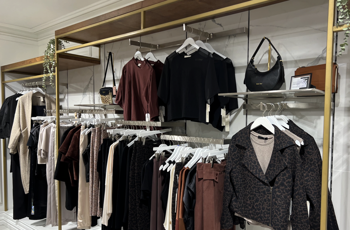 Clothing boutique interior display with various women's autumn apparel on gold clothing racks against marble walls.
