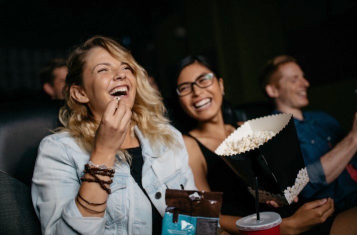 Three friends are seen enjoying a movie together, laughing and eating popcorn in a cinema theater.