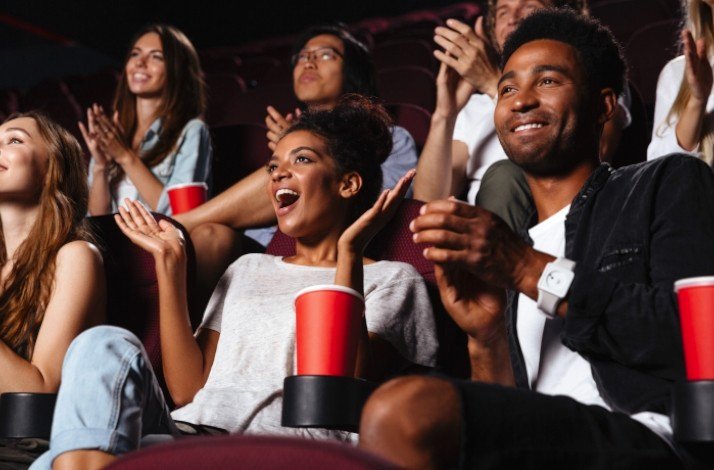 A group of people are seated, reacting with expressions of excitement and joy, in a cinema theater.