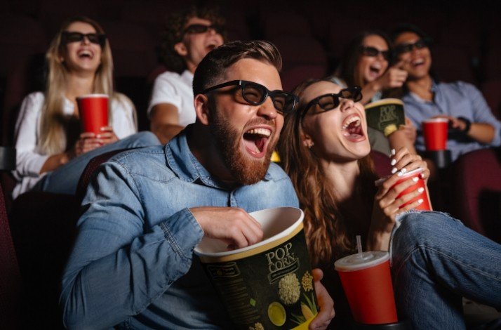 A group of people are wearing their 3d glasses whilst seated, laughing and eating popcorn in a cinema theater.