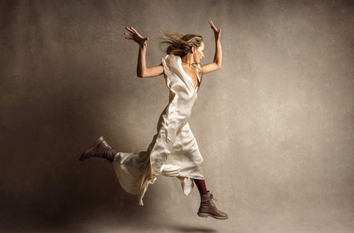 A Taryn Toomey in a light dress leaps into the air, showcasing her athleticism and energy against a clear background.