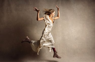 A Taryn Toomey in a light dress leaps into the air, showcasing her athleticism and energy against a clear background.