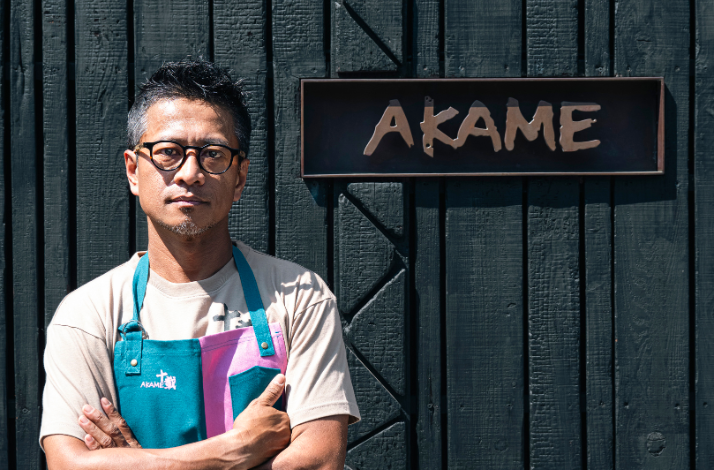 Chef Akame in apron standing in front of the Akame restaurant sign.