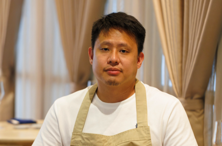 Chef Gen in a white shirt and beige apron seated in a softly lit dining room.