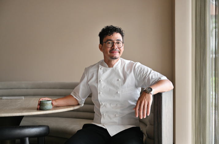 Chef Fiz in a white chef’s coat sitting at a table, holding a cup, in a modern restaurant.