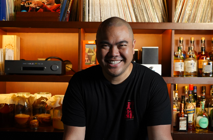 Man in a black shirt seated at a bar with shelves of bottles and records behind him.