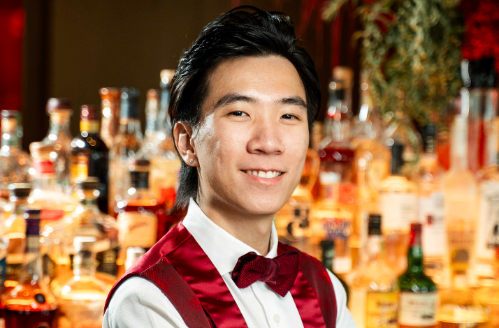 Bartender in a red vest and bow tie standing in front of a bar with bottles.