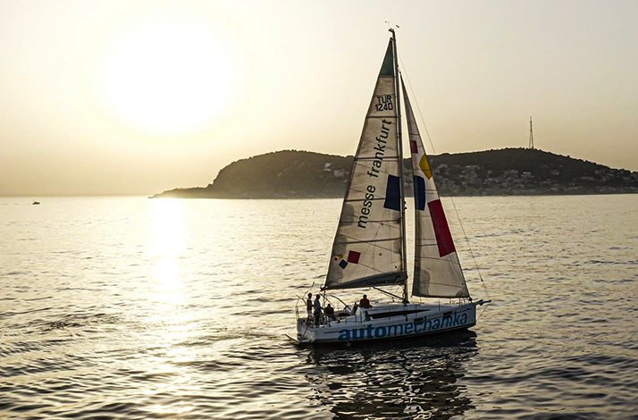 A sailboat with logos on its sails cruising on the sea during a golden sunset, with an island silhouette and sun reflections on the water in the background.