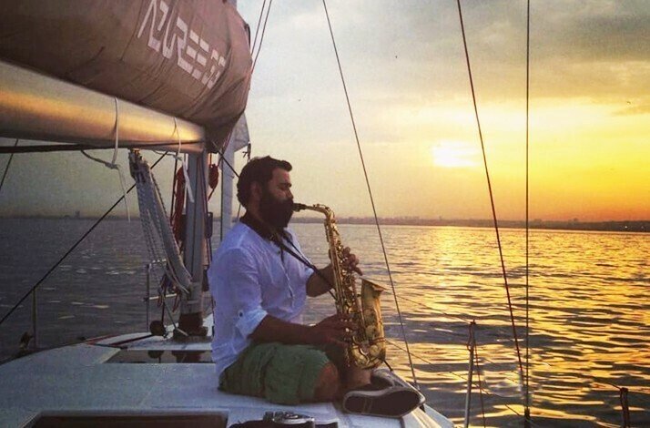 A bearded man in a white shirt sits on the deck of a sailboat in the middle of the sea at sunset, playing a saxophone.