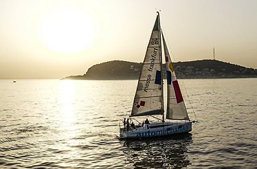 A sailboat with logos on its sails cruising on the sea during a golden sunset, with an island silhouette and sun reflections on the water in the background.