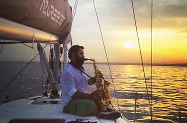 A bearded man in a white shirt sits on the deck of a sailboat in the middle of the sea at sunset, playing a saxophone.