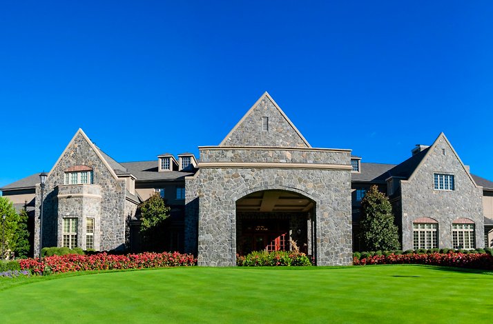 A large stone clubhouse with gabled roofs, an arched entrance, lush green lawn, and a clear blue sky at Alpine Country Club.