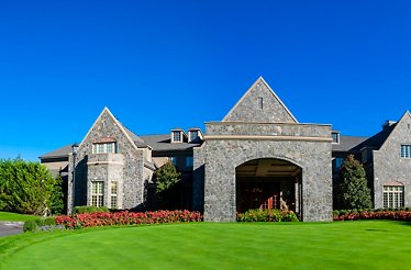 A large stone clubhouse with gabled roofs, an arched entrance, lush green lawn, and a clear blue sky at Alpine Country Club.