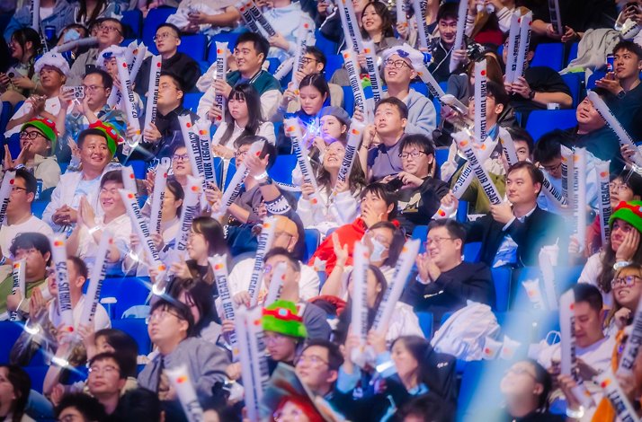Fans cheering at League of Legends Worlds 2024, waving Mastercard 'Make Them Believe' clappers, with 'No Jungler Pls' sign visible.