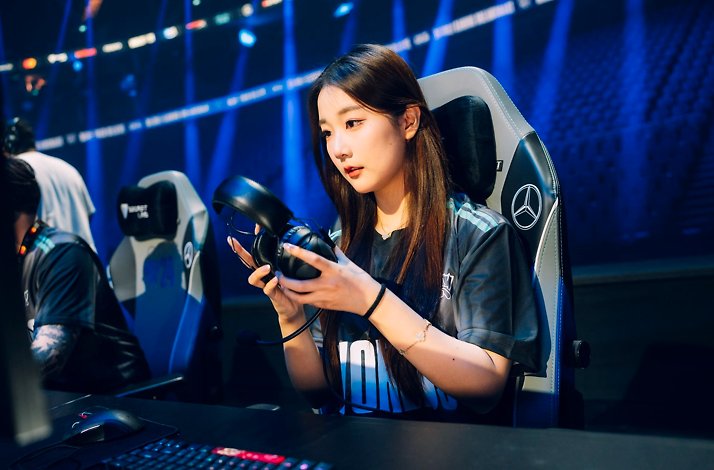 A player seated at a League of Legends Worlds pro stage setup, holding a gaming headset and wearing a team jersey.