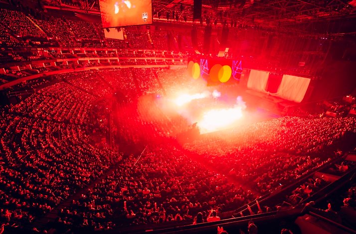 Sold-out arena lit in red with pyrotechnics blazing on stage at League of Legends Worlds; Logo of Mastercard displayed on center-rear screen.