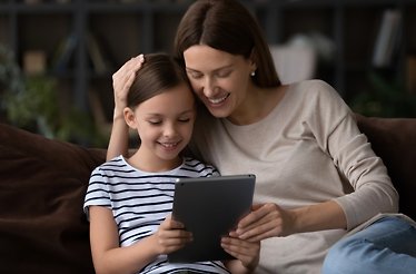 A child an a parent sitting on a couch looking at a tablet.