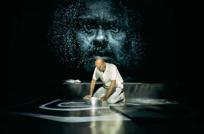 The performer kneels on stage beneath a large projected face.