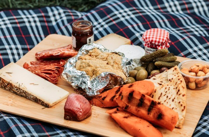 A close shot of the food set for a picnic on a checkered plaid - meat, cheese and  appetizers.